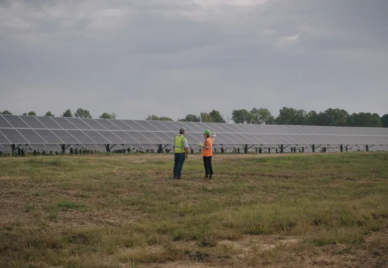Two people in safety gear stand in a grassy field near large rows of solar panels under a cloudy sky.