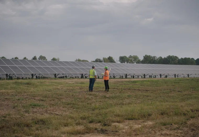 Two people in safety gear stand in a grassy field near large rows of solar panels under a cloudy sky.