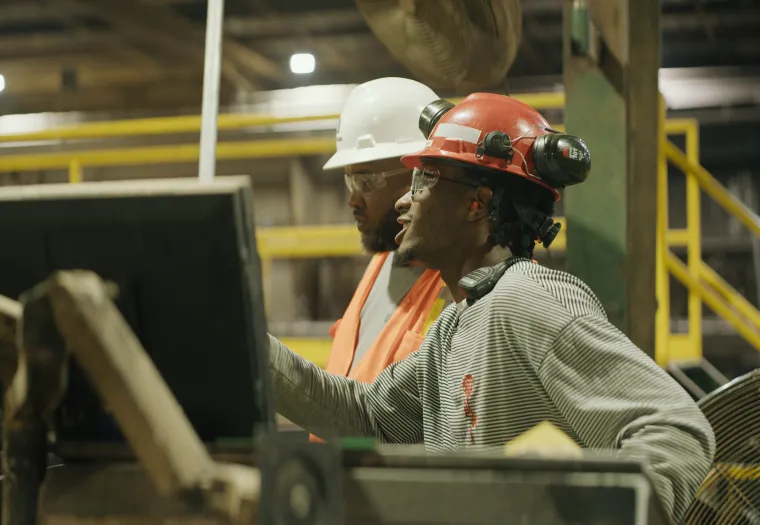 Two workers in safety gear operate machinery in an industrial setting, focusing on a computer monitor.
