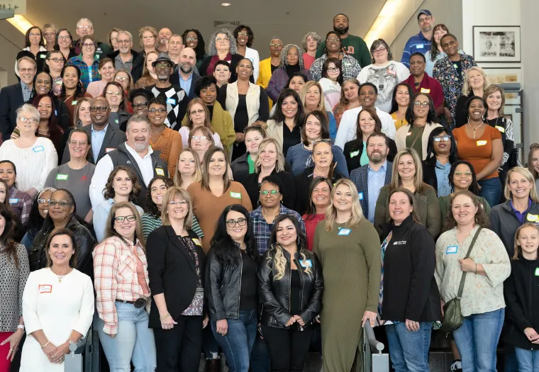 A large group of people stands on indoor stairs, smiling at the camera. Some individuals have name tags.