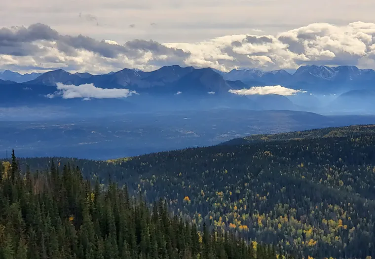 Mountainous landscape with layers of forest and hills under a cloudy sky. Distant peaks create a dramatic silhouette.