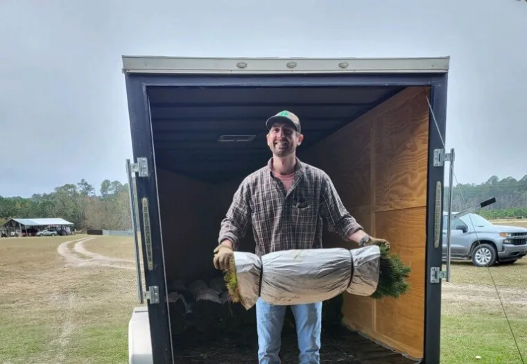 Man standing in a trailer holding a large wrapped plant, wearing a cap and plaid shirt.