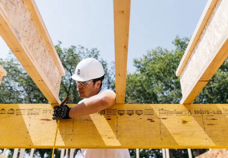 A construction worker wearing a white hard hat and safety glasses measures and marks a wooden beam at a construction site.
