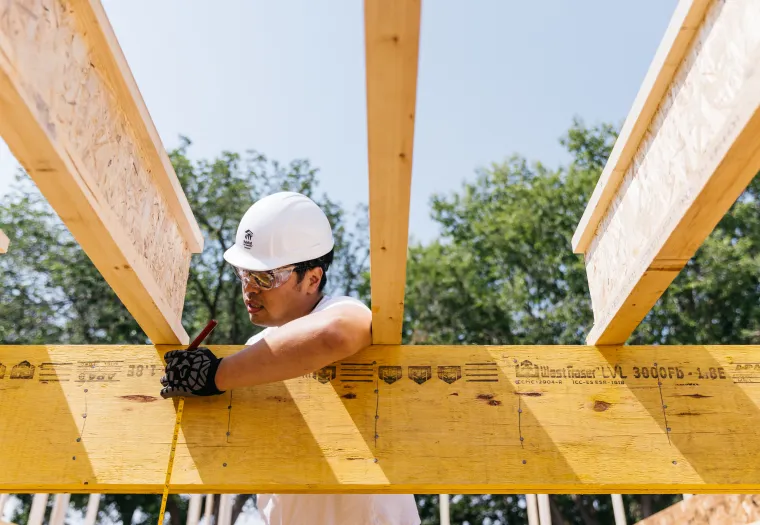 A construction worker wearing a white hard hat and safety glasses measures and marks a wooden beam at a construction site.