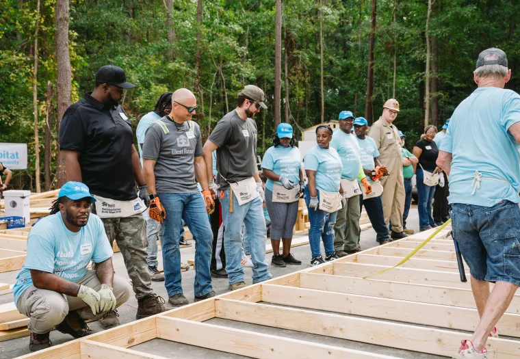 A group of people wearing casual work clothes and teal hats gather around wooden frames on the ground in an outdoor construction area.