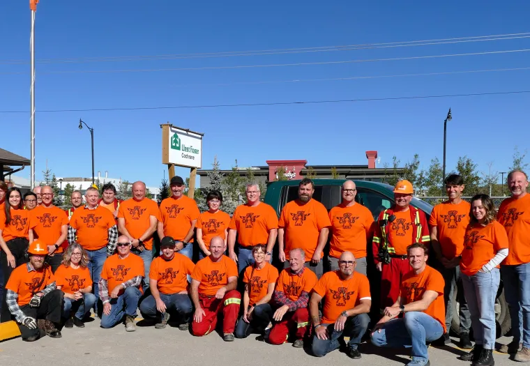 A group of people wearing orange shirts and outdoor gear pose for a photo in front of a building under a clear blue sky.
