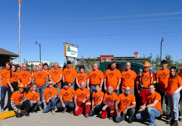 A group of people wearing orange shirts and outdoor gear pose for a photo in front of a building under a clear blue sky.