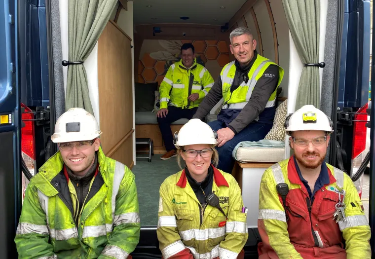 Four workers in safety gear smile for a group photo, seated in the back of a van with a wooden interior. Two additional people sit behind them.