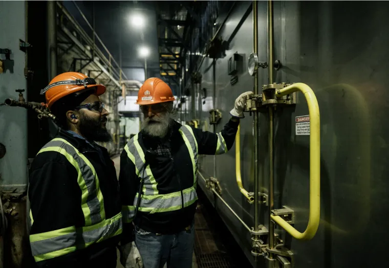 Two workers in safety gear inspect a large industrial machine inside a dimly lit facility.