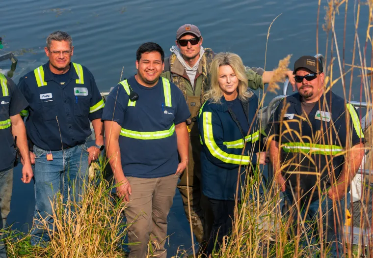 A group of six people wearing safety vests and work attire stand near water with tall grass in the foreground.