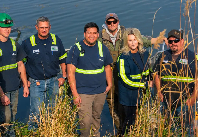A group of six people wearing safety vests and work attire stand near water with tall grass in the foreground.