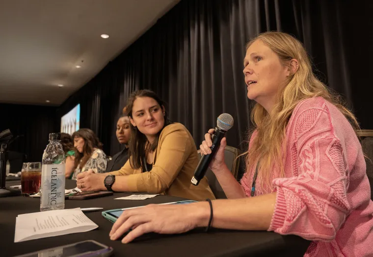 Four women sit at a conference table. One is speaking into a microphone. A water bottle and papers are on the table. Black curtains are in the background.