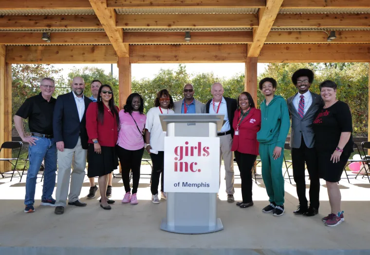 A group of people stands together under a wooden pavilion, with a podium labeled "Girls Inc. of Memphis" in front.