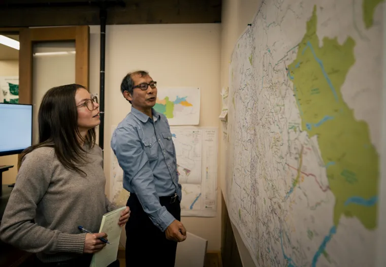 Two people examining a large map on a wall. The man points at the map while the woman takes notes. They are in a room with additional maps and a monitor in the background.