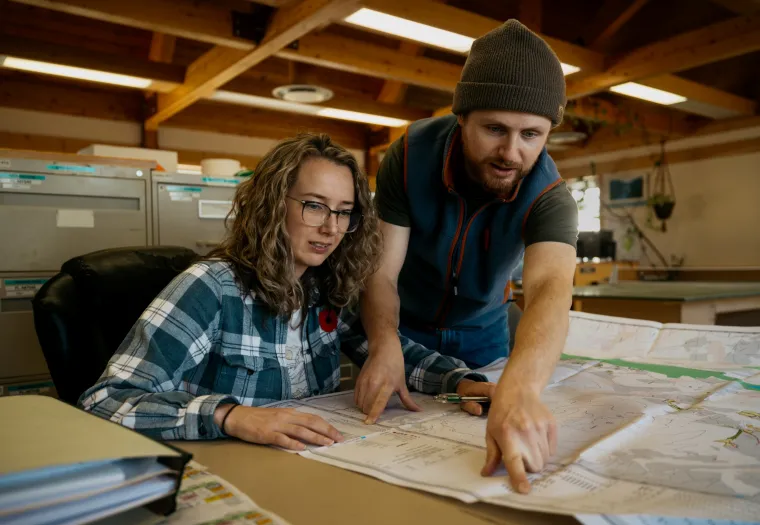 Two people reviewing large maps on a table. The woman is seated, wearing glasses, and the man is pointing at the map, wearing a beanie. They are in a room with wooden beams and filing cabinets.