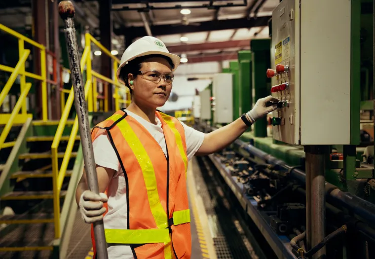 A person wearing a hard hat and reflective vest stands in a factory, holding a metal rod and adjusting controls on a panel.