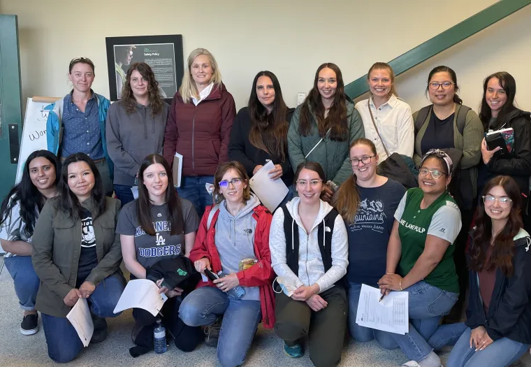 Group of people posing indoors, some holding papers. They stand and kneel in two rows. A staircase and a wall are in the background.
