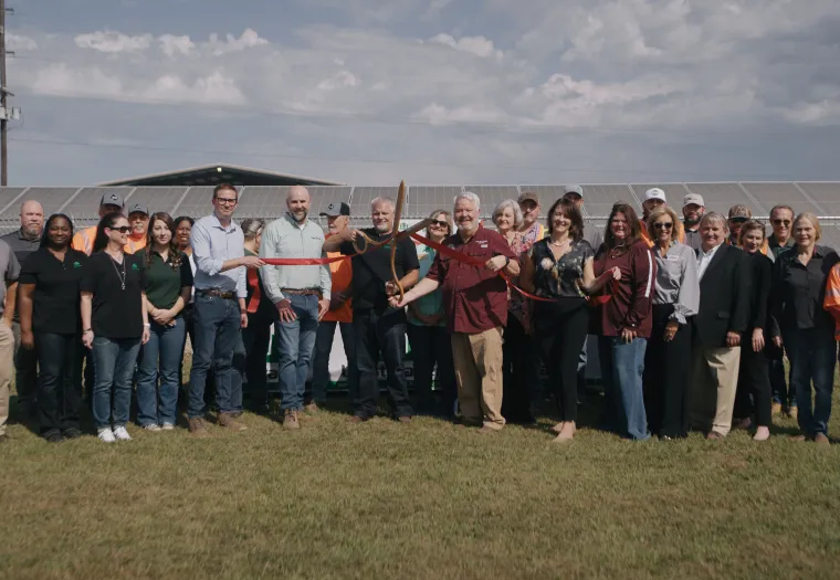 A group of people stand together outside in front of solar panels. One person is cutting a ribbon with large scissors. It is a sunny day with clouds in the sky.