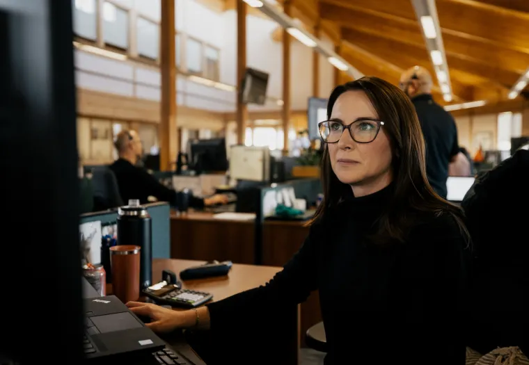 A woman with glasses works at a computer in an office with wooden ceilings. Other people are seen working in the background.