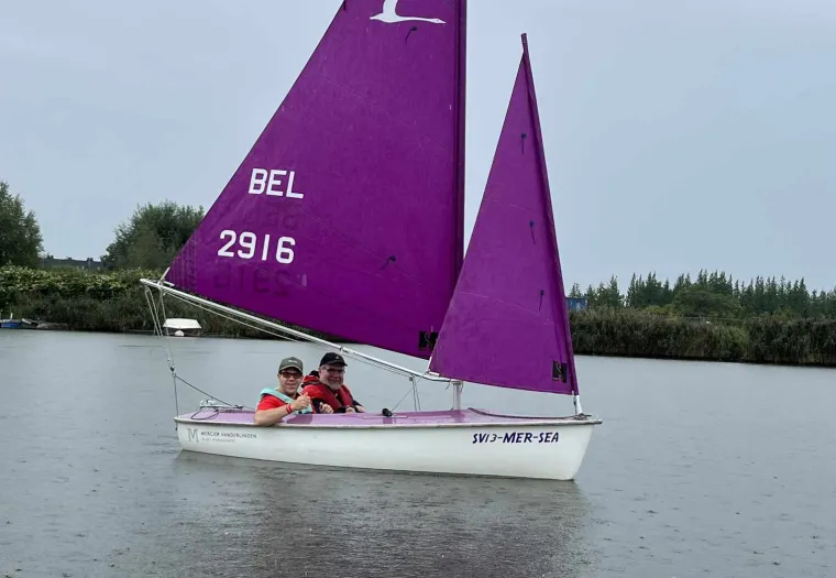 A small sailboat with a purple sail labeled "BEL 2916" floats on a calm body of water. Two people are seated inside, wearing life jackets. Trees line the background under a cloudy sky.