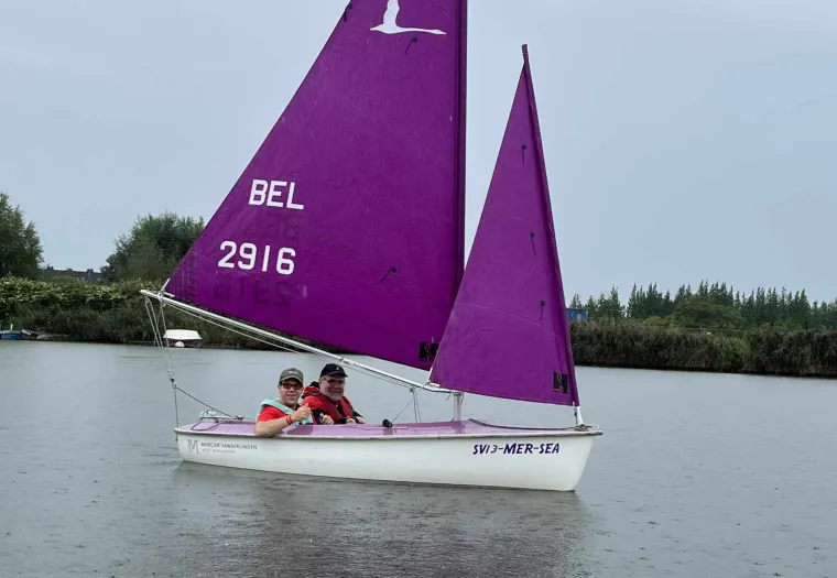 A small sailboat with a purple sail labeled "BEL 2916" floats on a calm body of water. Two people are seated inside, wearing life jackets. Trees line the background under a cloudy sky.