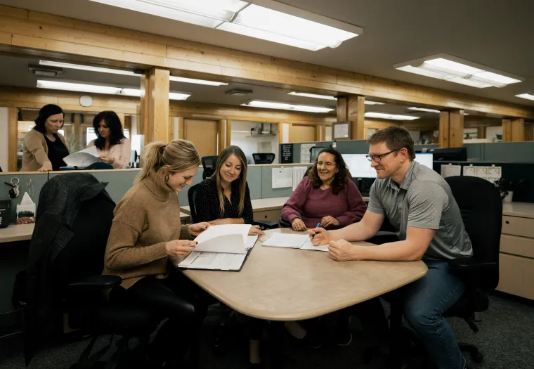 Four people sitting around a table in an office, discussing documents, with two others working in the background.