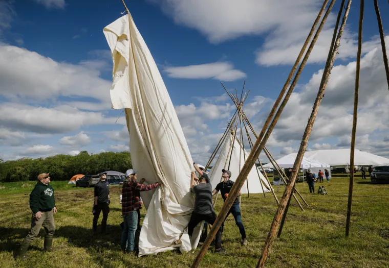 People are assembling a large canvas tent on a grassy field surrounded by poles under a partly cloudy sky.