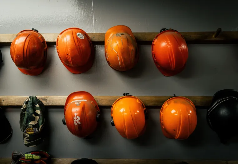 Six orange construction helmets and two other helmets, including a camouflage one, hang on a wooden rack against a gray wall.