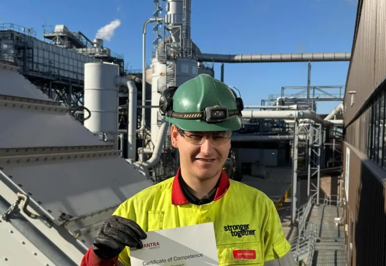 A worker wearing safety gear and holding a certificate stands in front of industrial equipment with smoke rising from a stack.
