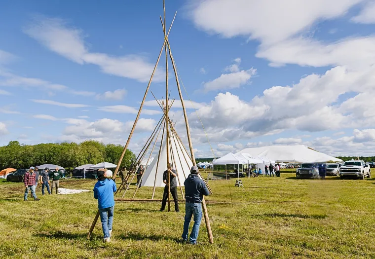 People are assembling a large teepee frame on a grassy field, with tents, parked vehicles, and cloudy blue skies in the background.
