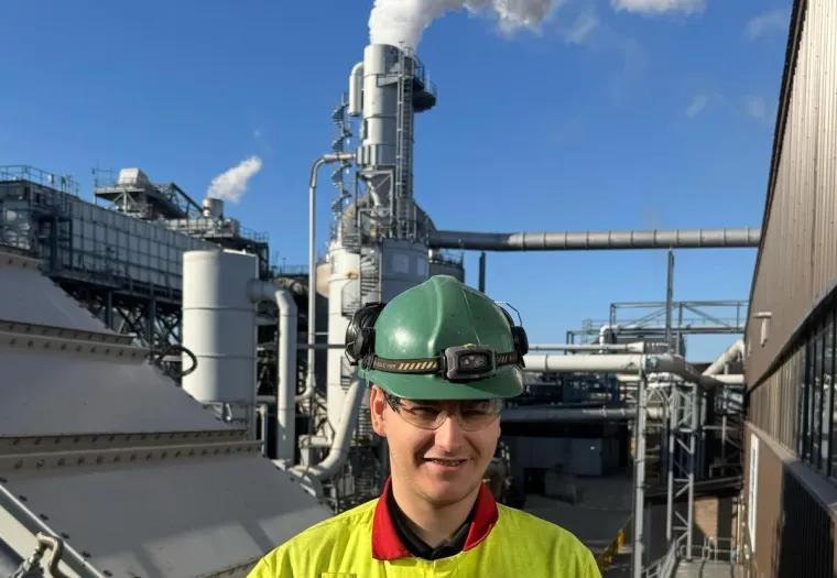 A worker wearing safety gear and holding a certificate stands in front of industrial equipment with smoke rising from a stack.