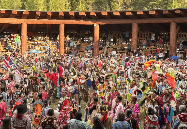 Large group of people in colorful traditional regalia participate in a powwow dance inside a wooden arbour, with spectators seated in the background.