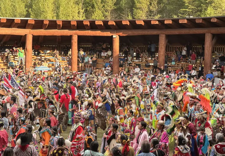 Large group of people in colorful traditional regalia participate in a powwow dance inside a wooden arbour, with spectators seated in the background.