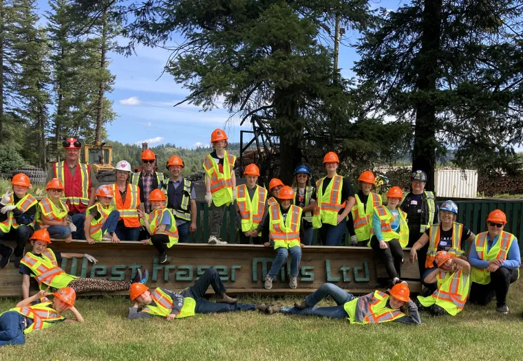 A group of kids wearing safety vests and hard hats pose together outdoors in front of a sign reading "West Fraser Mills Ltd." surrounded by trees.