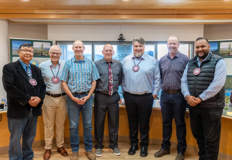 Seven men stand indoors in a row, dressed in business casual and formal attire, with meeting materials visible in the background.
