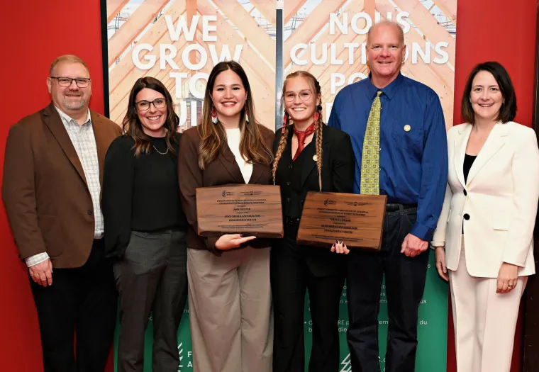 Six people stand indoors, with two women in the center holding award plaques. They are dressed in business attire and are smiling at the camera. Banners and a red wall are in the background.