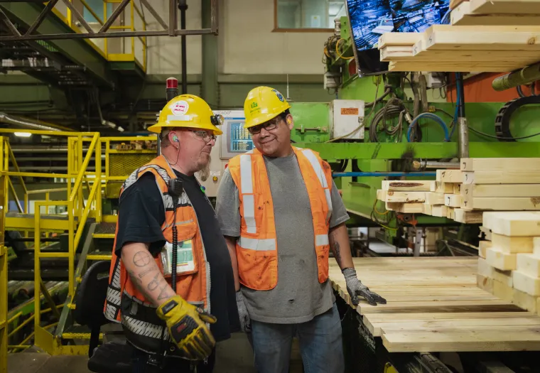 Two construction workers wearing hard hats and safety vests stand beside a stack of wooden planks inside an industrial facility, engaged in conversation.