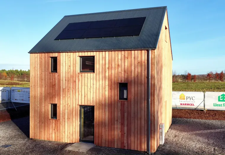 A two-story wooden house with a metal roof and solar panels, surrounded by construction barriers on a gravel lot in a rural area.