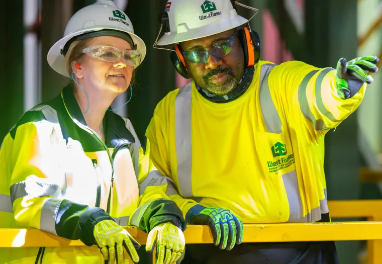 Two mill workers in safety gear stand by a yellow railing; one points ahead while the other listens attentively.