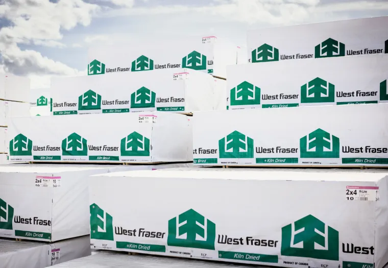 Stacks of kiln-dried lumber wrapped in white packaging with West Fraser logo and branding under a partly cloudy sky.