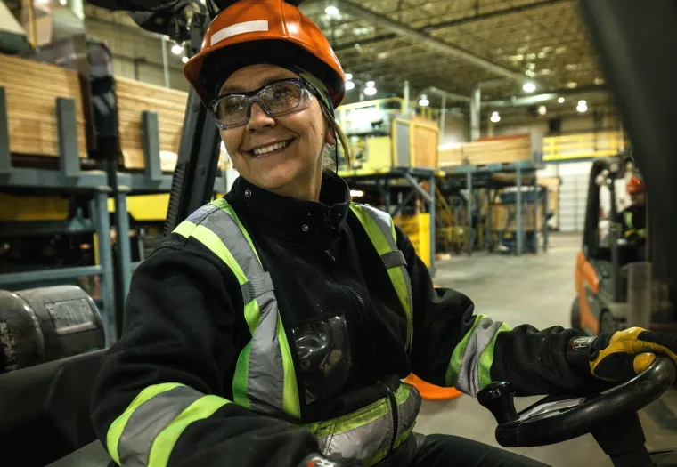 Person wearing a hard hat, safety glasses, and reflective jacket operates machinery inside an industrial warehouse.