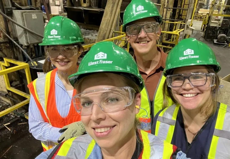 Four people wearing green West Fraser hard hats, safety glasses, and reflective vests pose for a selfie inside an industrial facility.
