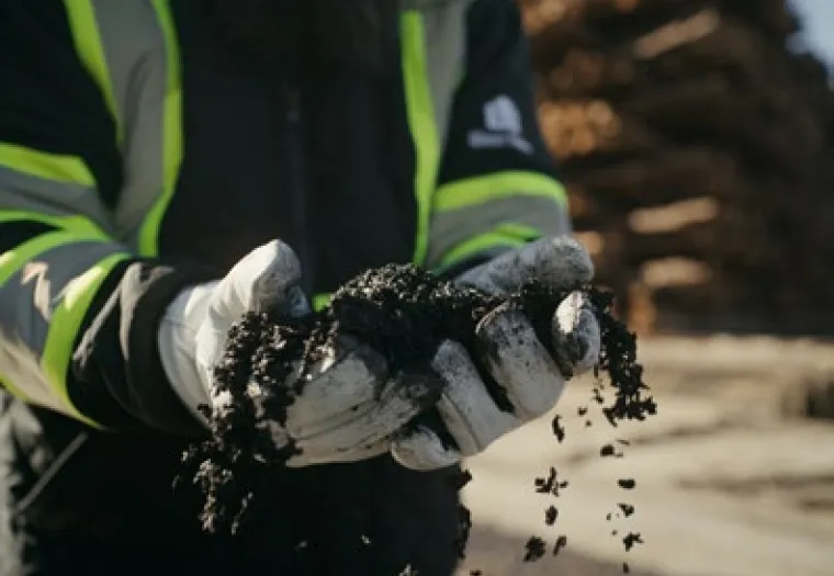 A person wearing gloves and a reflective jacket holds a handful of biochar outdoors, with stacked logs blurred in the background.
