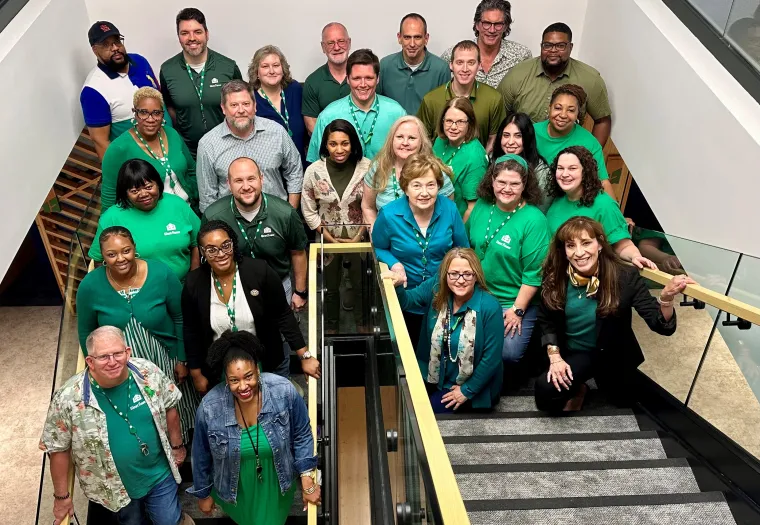 A group of people, many wearing green, stand and sit on a staircase, posing for a group photo in an indoor setting.