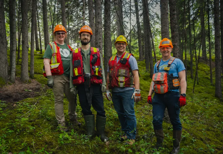 Four people wearing safety vests, helmets, and work gloves stand together in a forested area with green undergrowth and trees in the background.