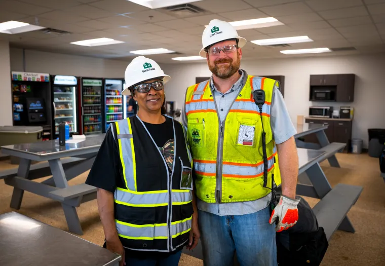Two construction workers in safety vests and hard hats stand together and smile in a break room with picnic tables, vending machines, and a kitchenette in the background.