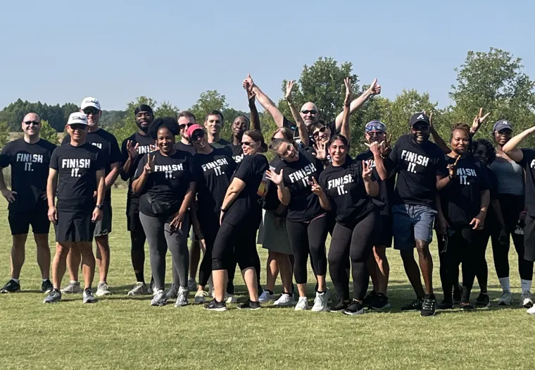 A group of people pose outdoors on grass, many wearing matching "Finish It." t-shirts, with trees and a lake in the background under a clear sky.