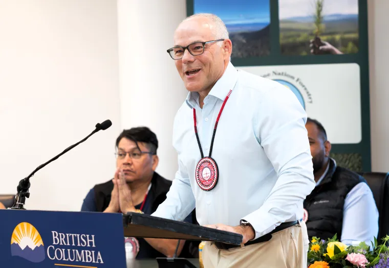 A man wearing glasses and a medallion speaks at a podium with a British Columbia sign; two people sit behind him, one with hands clasped, and a floral arrangement is nearby.