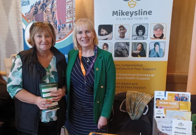 Two women stand smiling at a Mikeysline mental health support booth, featuring informational leaflets, brochures, and display banners.