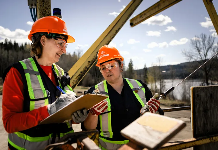 Two workers in safety vests and helmets stand outdoors, one holding a clipboard and pen, the other holding a tool, with trees and water in the background.